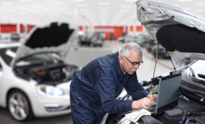 a photo of an engineer looking into tapping on the power of big data and transportation as he works on his laptop sitting on top of an open hood of a car's engine in an auto shop