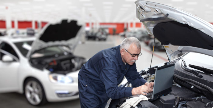 a photo of an engineer looking into tapping on the power of big data and transportation as he works on his laptop sitting on top of an open hood of a car's engine in an auto shop