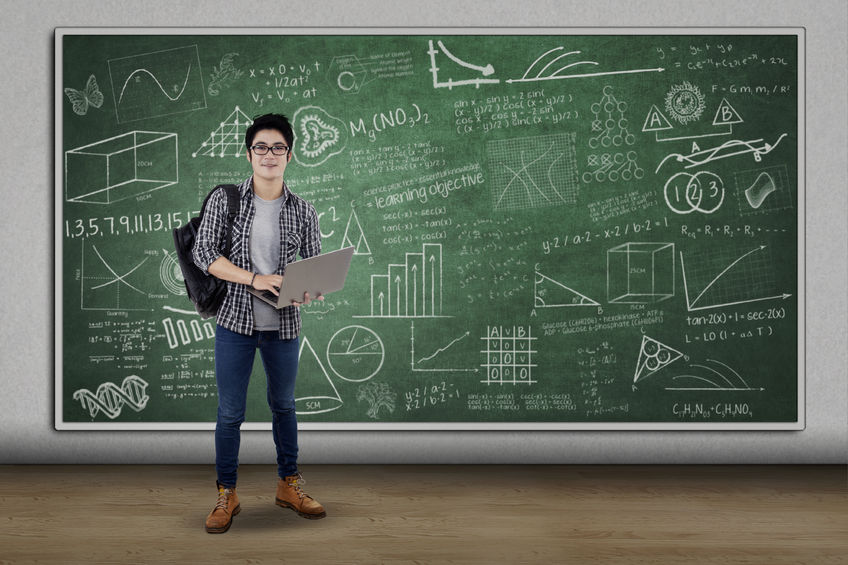 a photo of a young man holding a laptop and carrying a backpack stands in front of green chalkboard at one of the charter schools in NYC