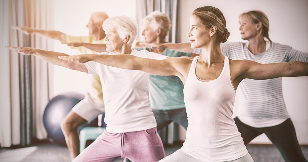 a photo of elderly men and women doing yoga stretches as instructed by a young woman in a class amid discussions about healthcare for senior citizens