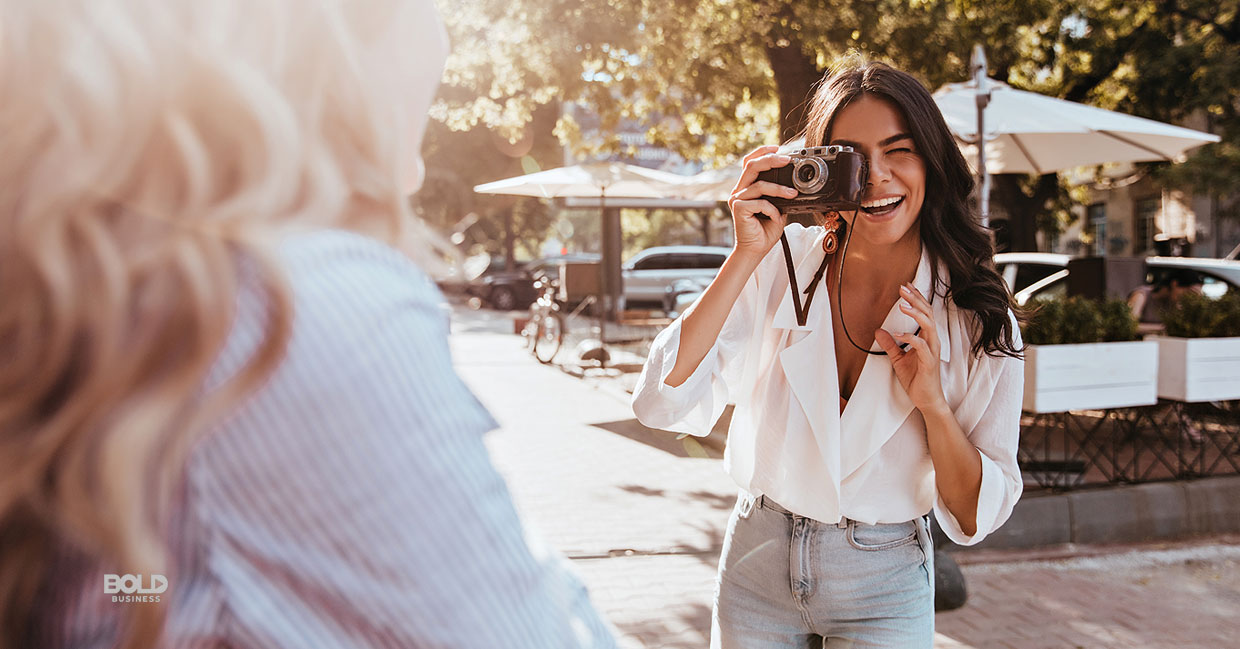 a photo of a young brunette woman looking through a camera and taking a photo of a young blonde woman while they're standing on the sidewalk of a street amid the bold impact of Girlgaze